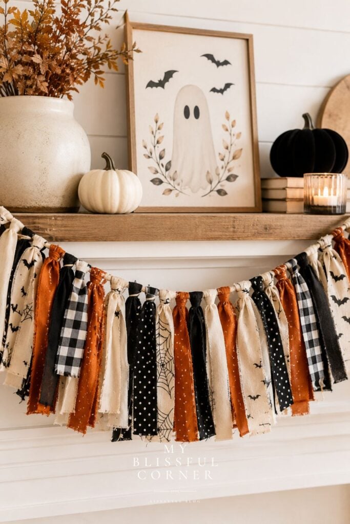 DIY Halloween rag garland hanging on a farmhouse mantel with neutral Halloween decor, pumpkins, ghost artwork, and cozy fall accents.