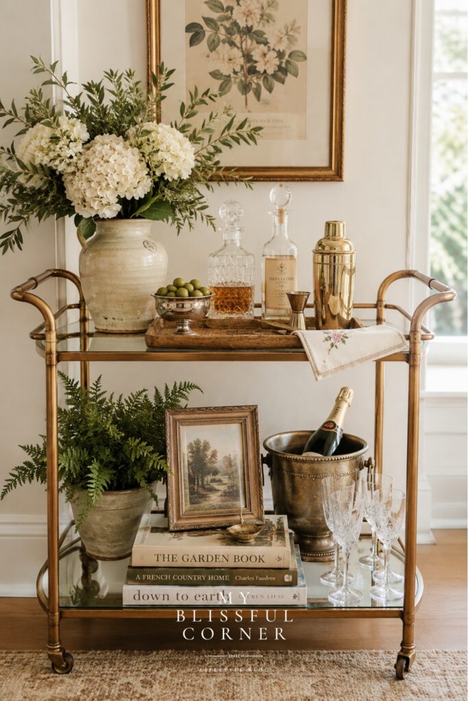 Neutral bar cart styling with layered decor, fresh greenery, vintage artwork, champagne bucket, and warm organic textures in a cozy home corner.