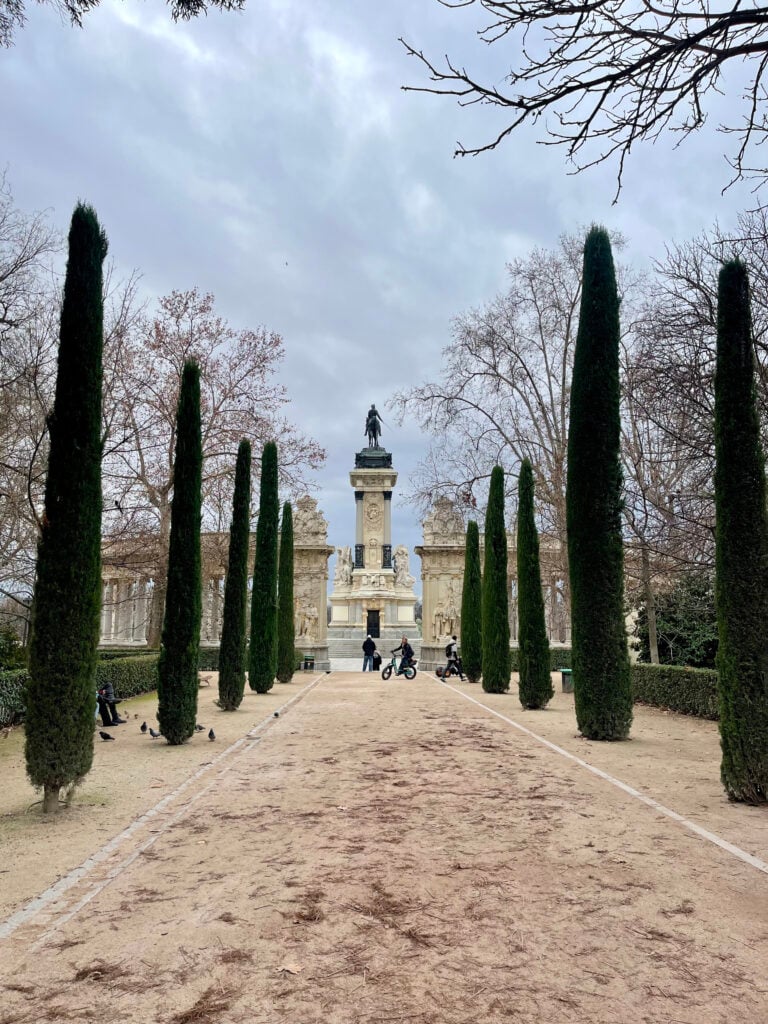 Tree-lined walkway in Retiro Park Madrid leading toward a statue under cloudy skies