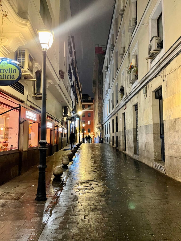 Narrow historic street in Madrid at night with warm lights reflecting on wet stone pavement