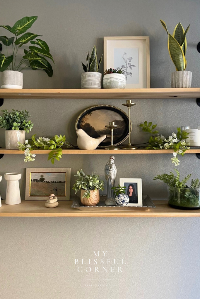Living room shelf decor with natural wood floating shelves styled with greenery, framed art, brass candle holders, ceramic vases, and personal photos.