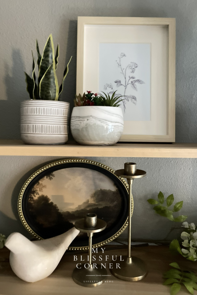 Living room shelf decor with wood floating shelves styled with potted plants, framed botanical art, brass candle holders, and layered decorative accents.