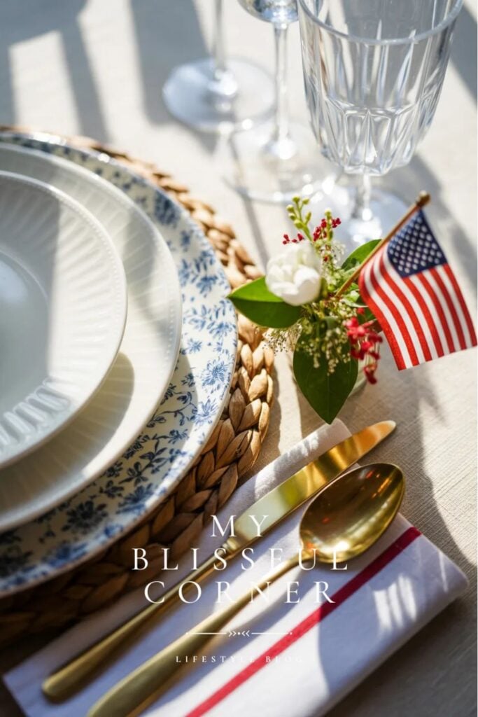 Patriotic table setting with blue and white plates, gold flatware, woven placemat, and small American flag centerpiece