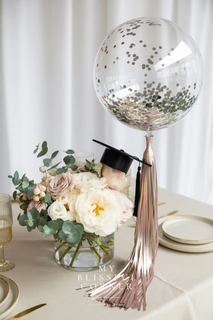 Elegant graduation centerpiece with clear confetti balloon, blush tassel, white flowers, greenery, and graduation cap decor