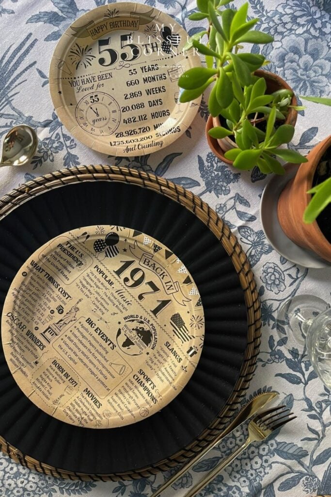 55th birthday table set up for men with black charger plates, wicker placemats, vintage newspaper themed 55th birthday disposable plates on a blue floral tablecloth