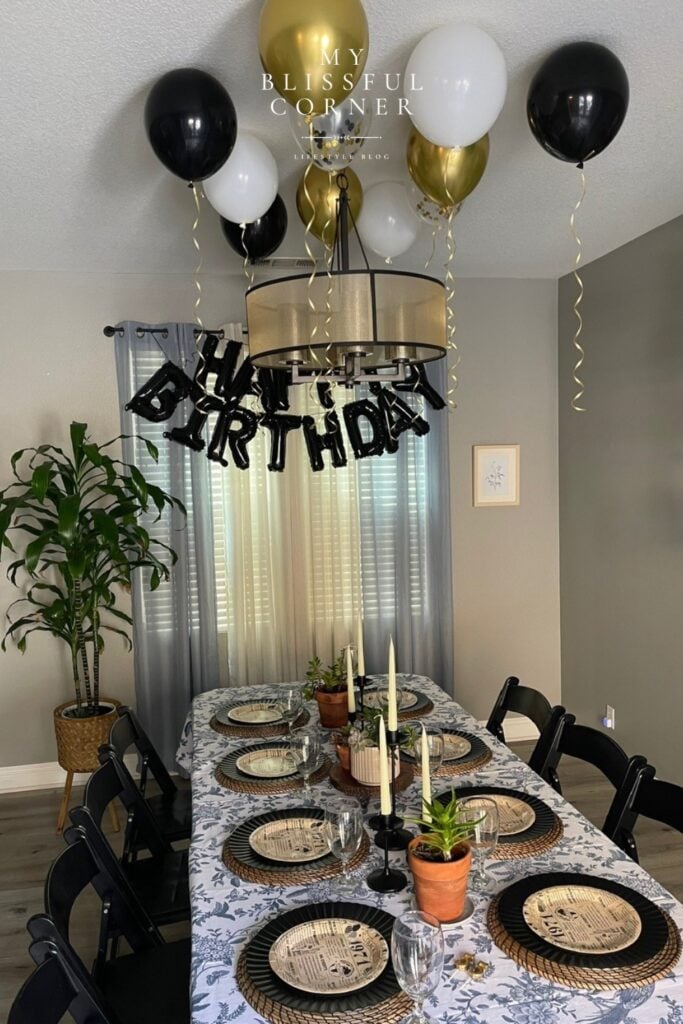 Birthday set up ideas for him at home with black and gold ceiling balloons, a happy birthday banner, and an elevated dinner table with wicker placemats and candlestick holders