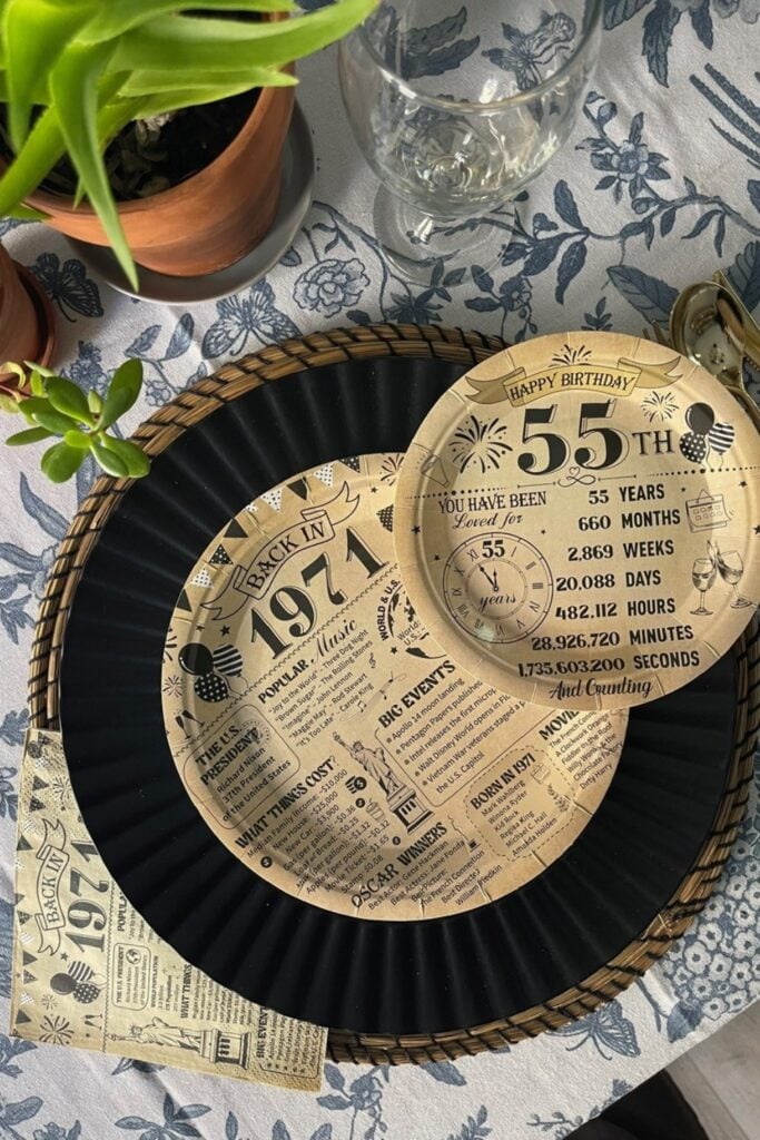 55th birthday table set up for men with black charger plates, wicker placemats, vintage newspaper themed 55th birthday disposable plates on a blue floral tablecloth
