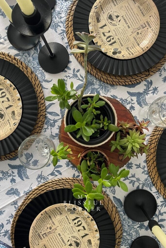 Men's birthday table set up with black charger plates, wicker placemats, succulents, black candlestick holders, and vintage newspaper themed disposable plates on a blue floral tablecloth