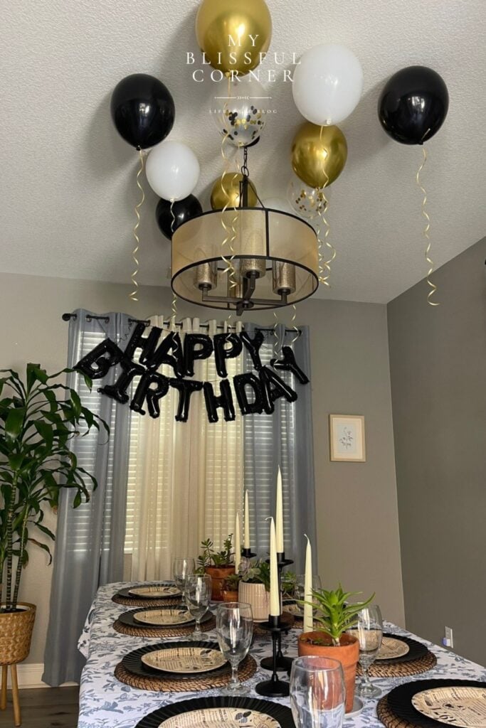 Men's birthday decorations at home with black Happy Birthday letter balloons as a photo backdrop, black white and gold ceiling balloons with hanging strings, and an elevated tablescape with wicker placemats and candlestick holders