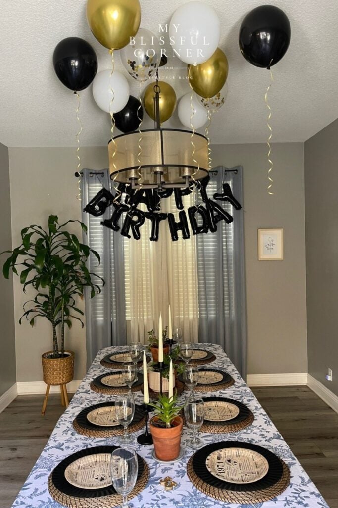 black white and gold ceiling balloons, black Happy Birthday letter balloons, and a simple birthday table with wicker placemats, candles and succulents on a blue floral tablecloth