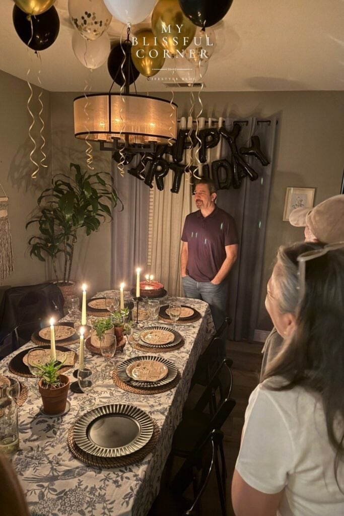 Hubby birthday party with an elevated dinner table, candles, black and gold ceiling balloons, and a black Happy Birthday letter balloon backdrop