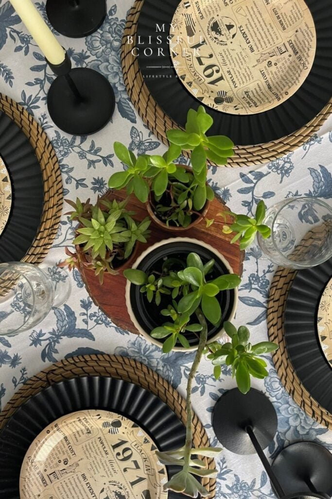 Men's birthday table set up with black charger plates, wicker placemats, succulents, black candlestick holders and vintage newspaper themed disposable plates on a blue floral tablecloth