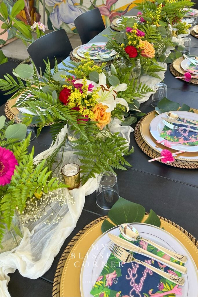 Tropical floral centerpiece styled on party table with luau place settings and greenery runner
