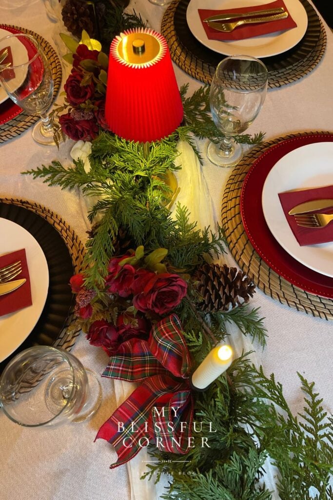 Traditional Christmas table with burgundy flowers, evergreen greenery, tartan ribbon, pinecones, woven chargers, and red place settings.