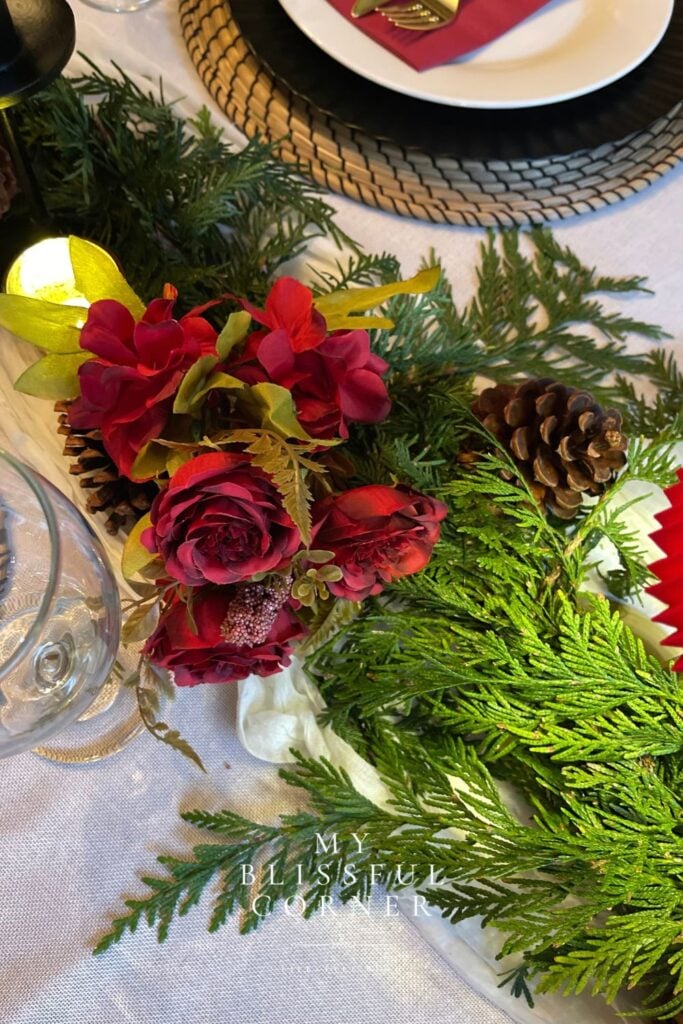 Close-up of an elegant Christmas centerpiece with deep-red flowers, plaid ribbon, evergreen branches, pinecones, and crystal glassware.
