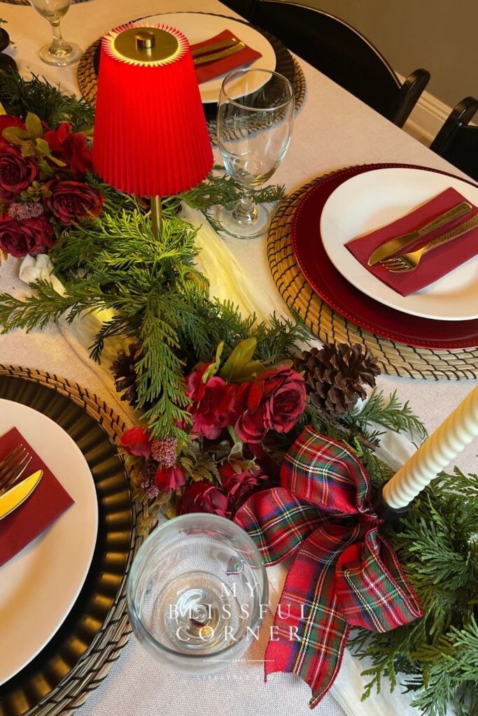 Layered Christmas place setting with a woven charger, white plate, burgundy napkin, gold flatware, and evergreen table runner.