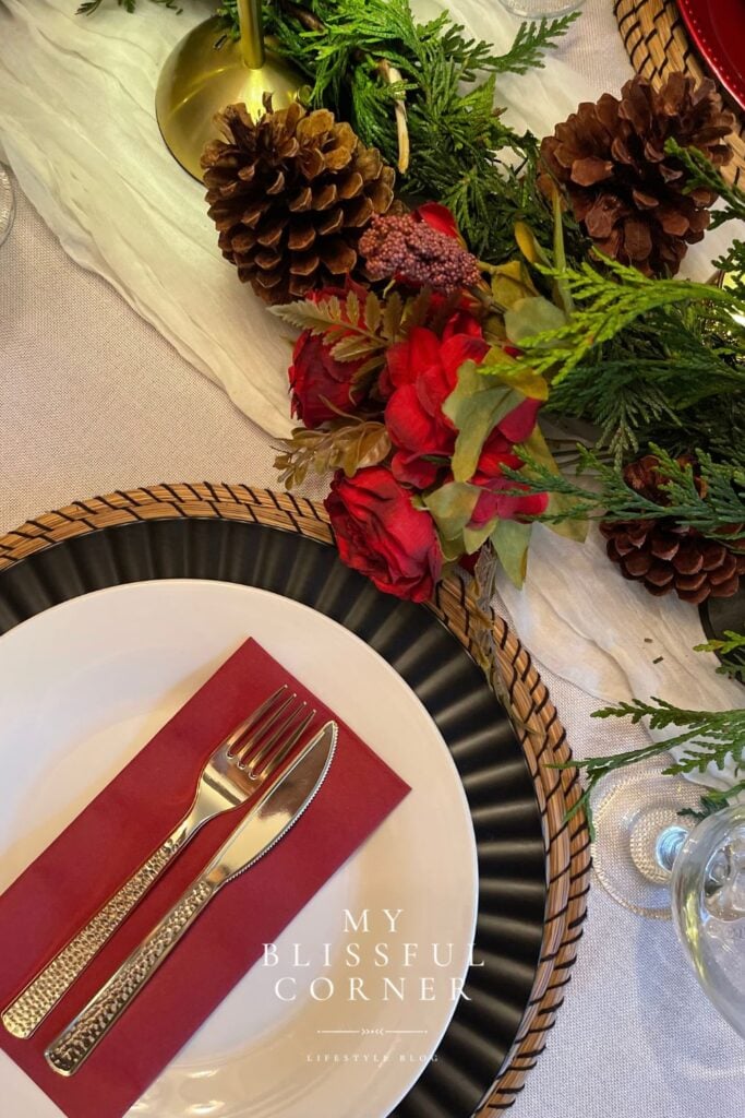 Overhead view of a Christmas table setup with evergreen garland, burgundy florals, tartan ribbon, red lamps, and layered place settings.
