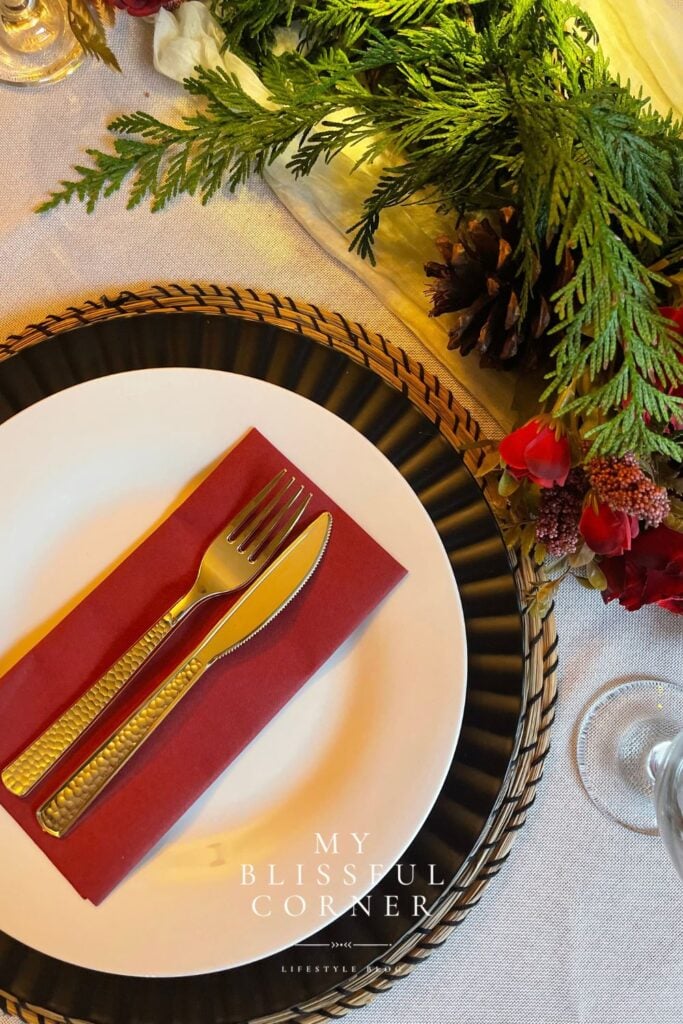 Evergreen Christmas table runner decorated with pinecones, burgundy flowers, and a red napkin layered over neutral dinnerware.