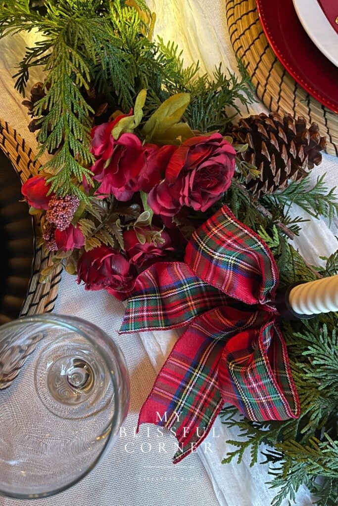 Evergreen Christmas table runner decorated with pinecones, burgundy flowers, and a red napkin layered over neutral dinnerware.