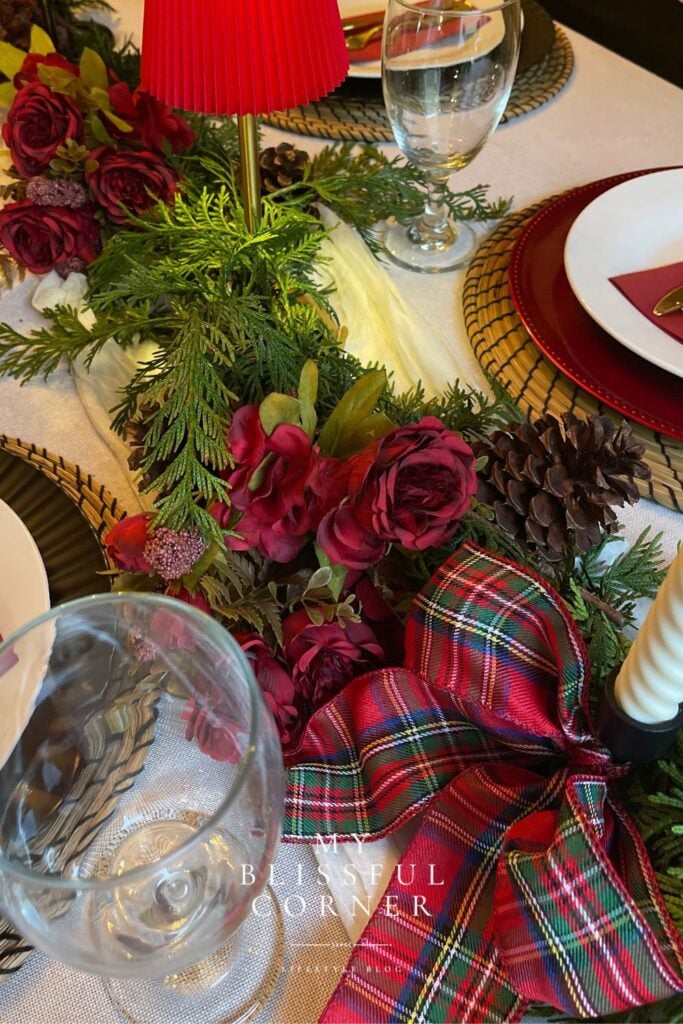 Candlelit Christmas dinner table with a red accent lamp, evergreen garland, deep-red flowers, plaid ribbon, and layered place settings.