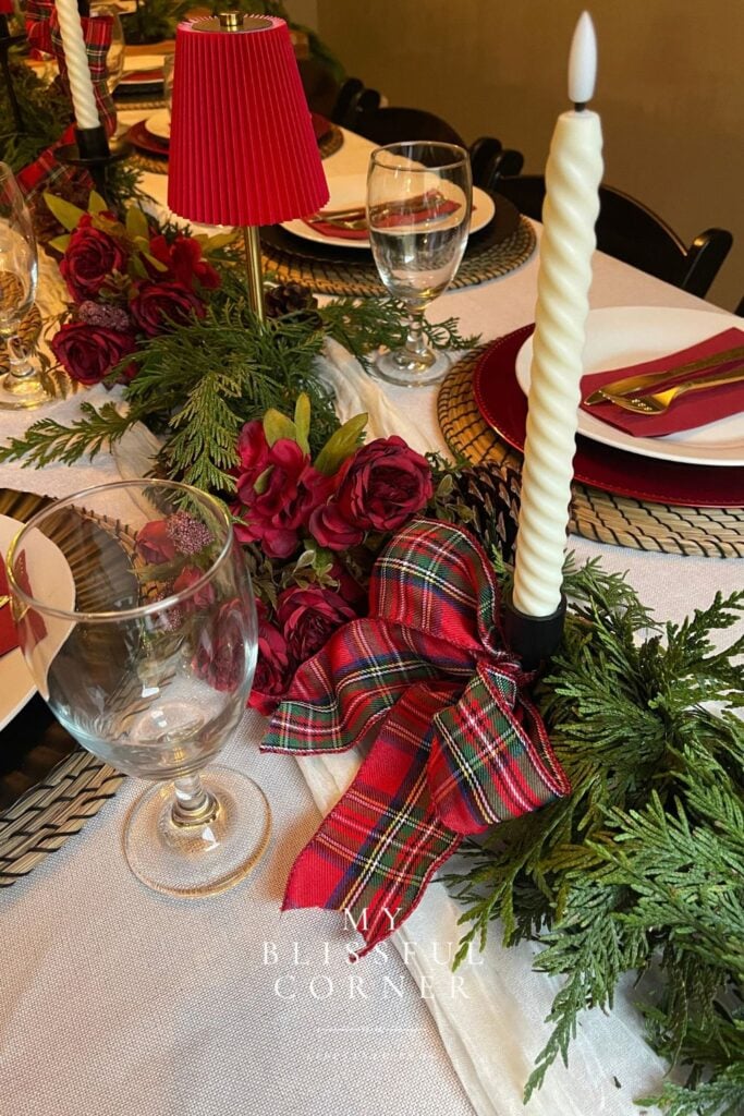 Deep-red floral Christmas centerpiece arranged with cedar greenery, pinecones, and woven place settings.