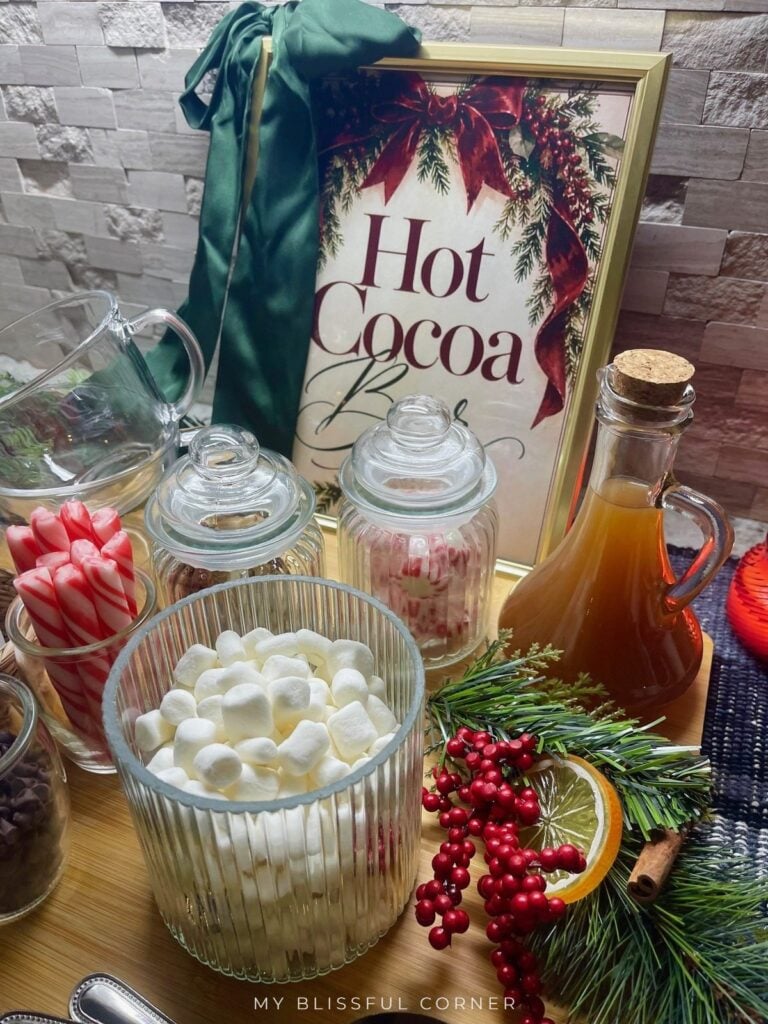 Close-up of a DIY hot cocoa bar with a ribbed glass jar of mini marshmallows, peppermint candy jar, caramel syrup, and red berries