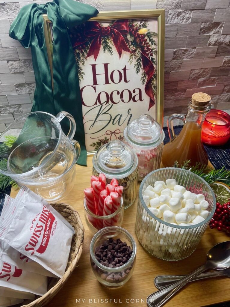Dollar Tree hot cocoa bar on a bamboo board with a framed Hot Cocoa Bar sign, glass mugs, jars of marshmallows and peppermint, and a green satin bow