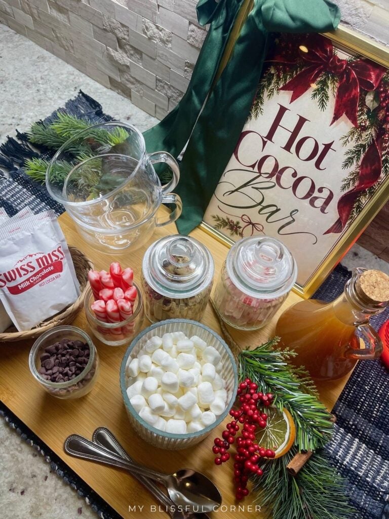 Overhead view of a DIY hot cocoa bar with mini marshmallows, peppermint sticks, chocolate chips, cocoa packets in a basket, and caramel syrup