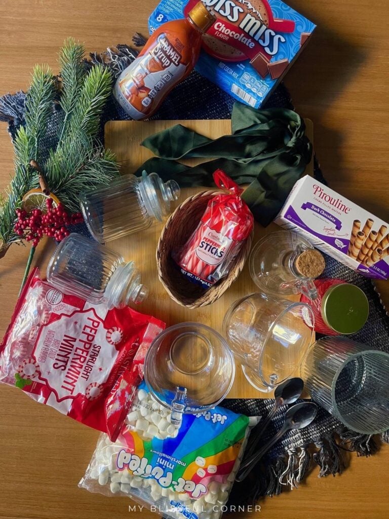 Dollar Tree hot cocoa bar supplies laid out on a bamboo board, including glass jars, glass mugs, a woven basket, peppermint candy, mini marshmallows, and cocoa mix