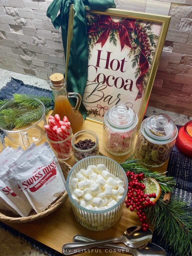 Hot cocoa bar on a budget with peppermint sticks, chocolate chips, a jar of chocolate wafer rolls, and a framed Christmas sign